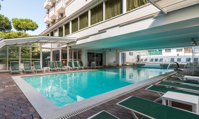 Piscina coperta in un hotel con lettini verdi e tavolini bianchi.