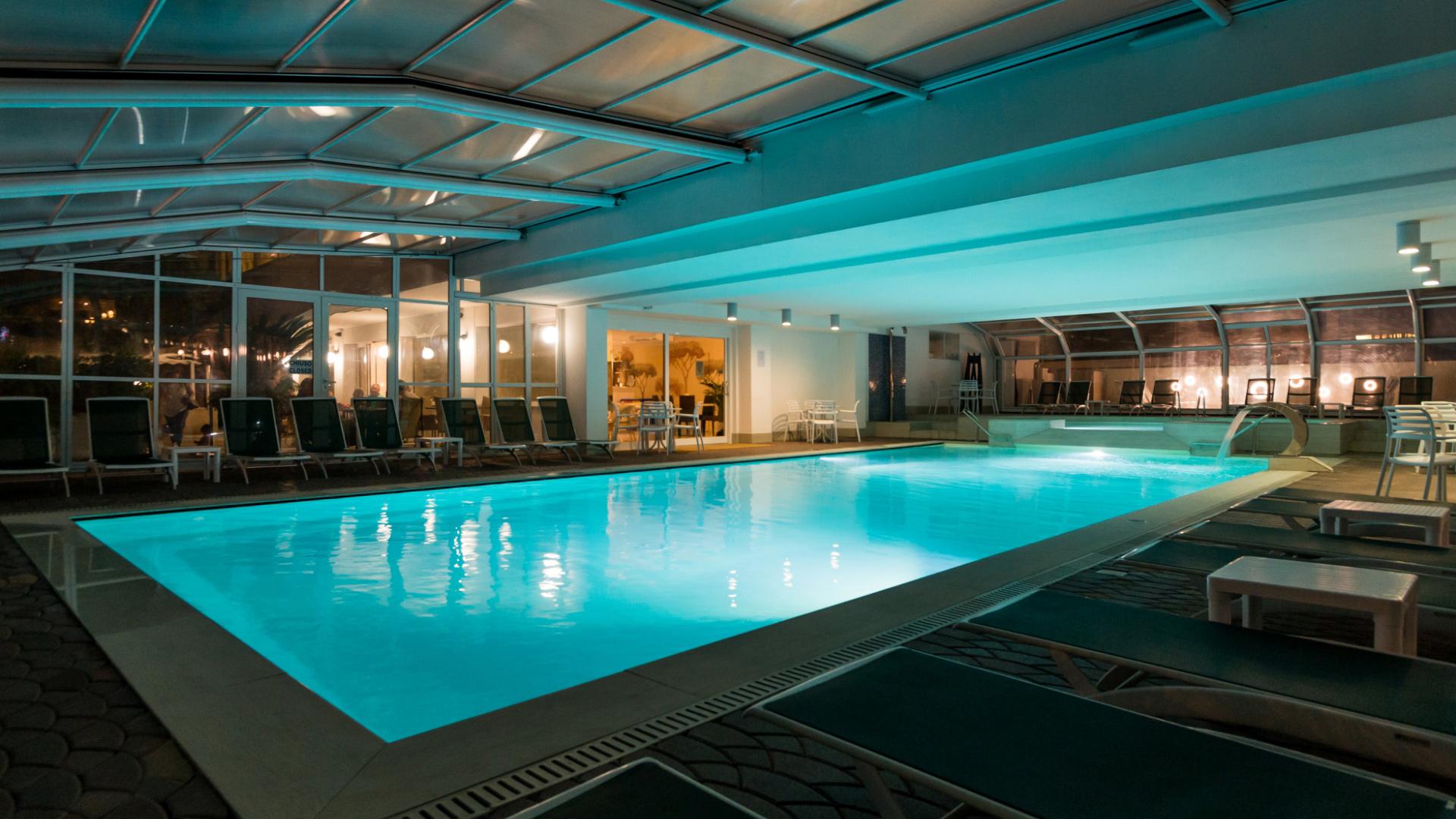 Piscina coperta illuminata di notte con lettini e area relax circostante.