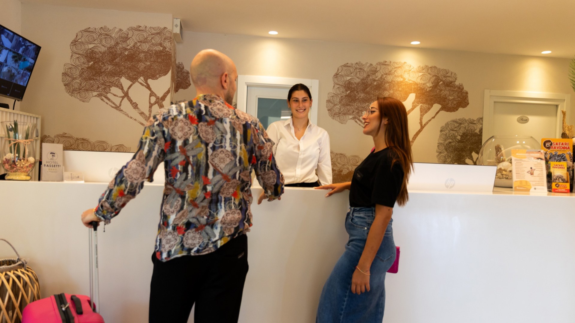 Persone alla reception di un hotel, conversano con una receptionist sorridente.