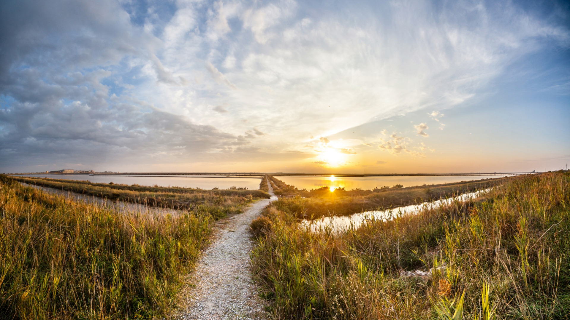 Sentiero tra le saline al tramonto, cielo nuvoloso e riflessi sull'acqua.