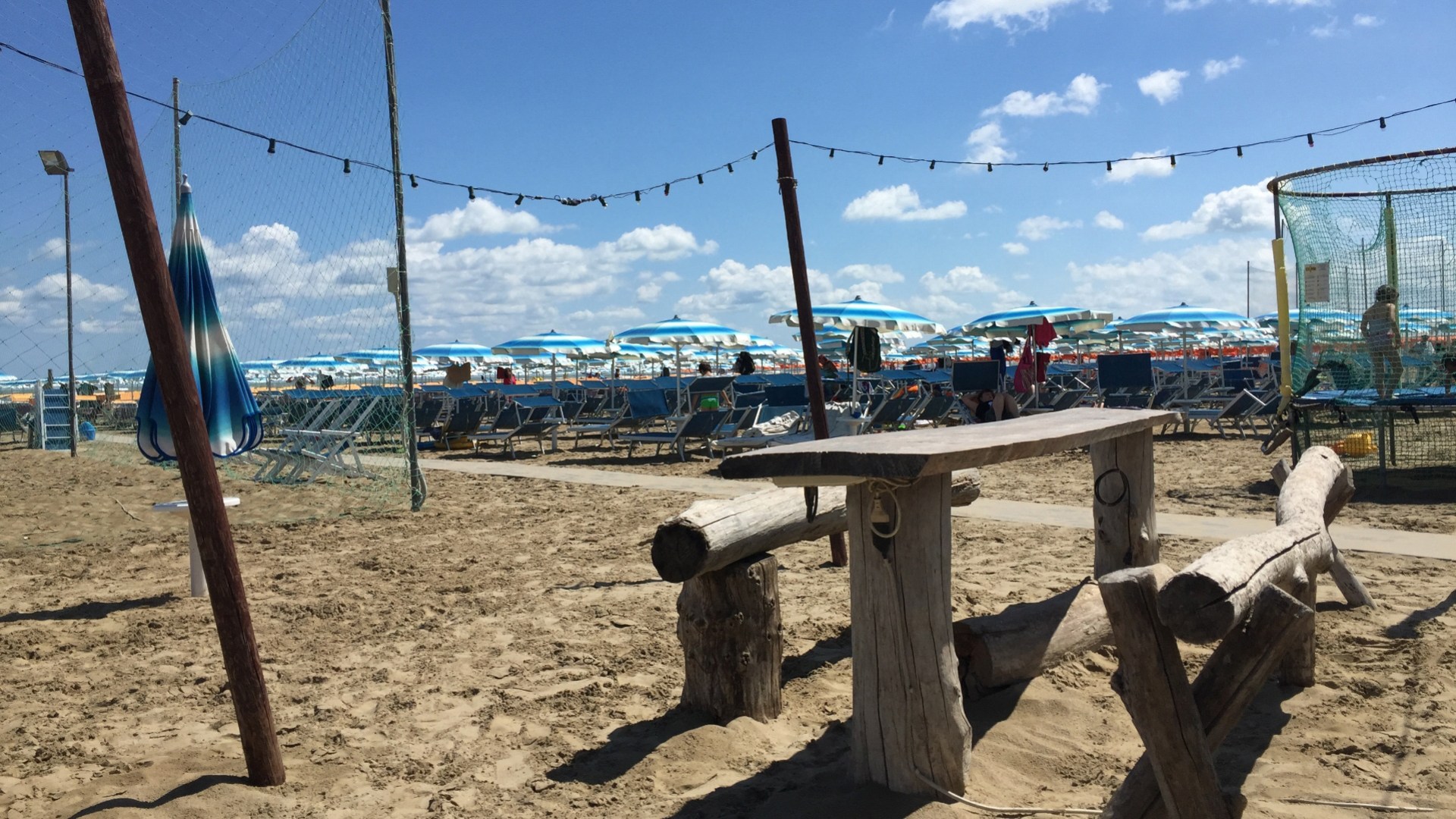 Spiaggia con ombrelloni blu e bianchi, tavolo rustico e sabbia soleggiata.