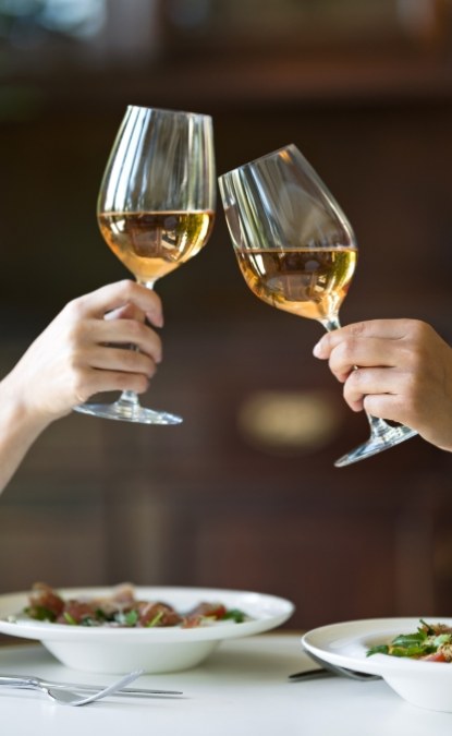 Due persone brindano con bicchieri di vino bianco sopra un tavolo apparecchiato.