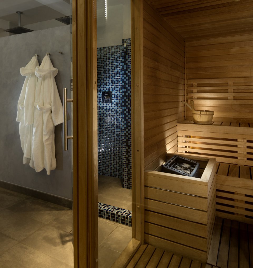 Sauna in legno con doccia piastrellata e accappatoi bianchi appesi.