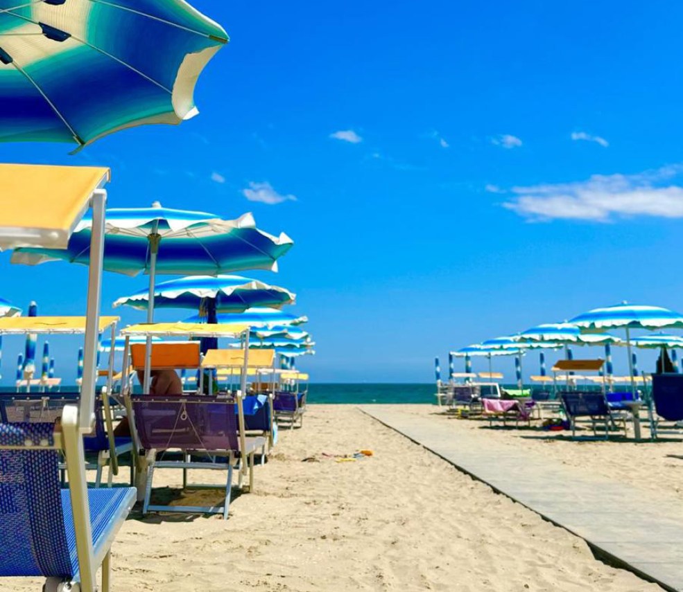 Bambino in spiaggia con cappello, occhiali da sole e costume a righe rosse.