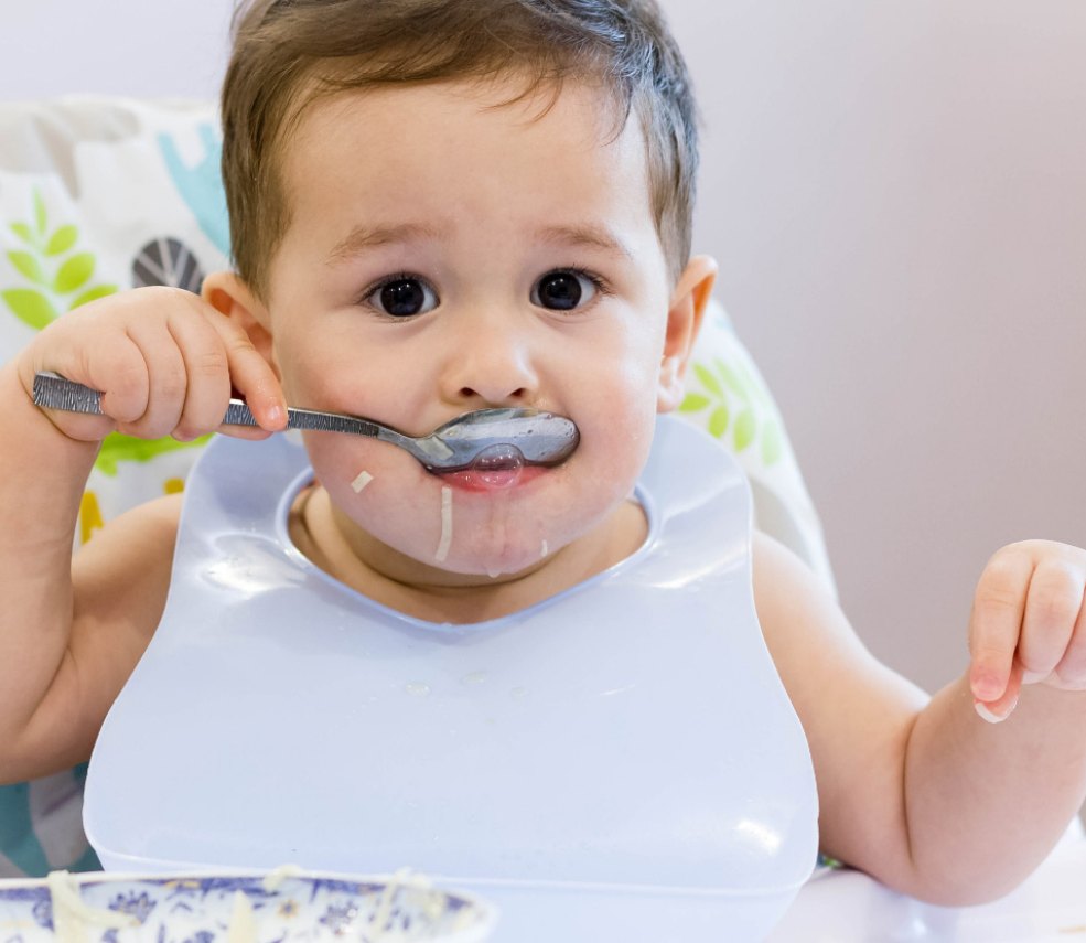 Bambino con bavaglino mangia con cucchiaio, seduto a tavola.